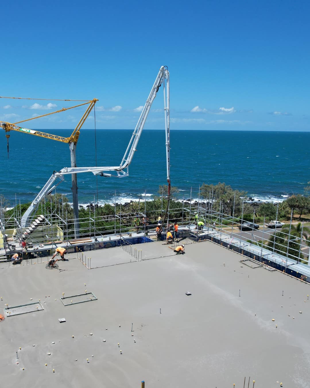 A concrete pump boom arcs over the freshly poured top floor, the Coral Sea filling the horizon