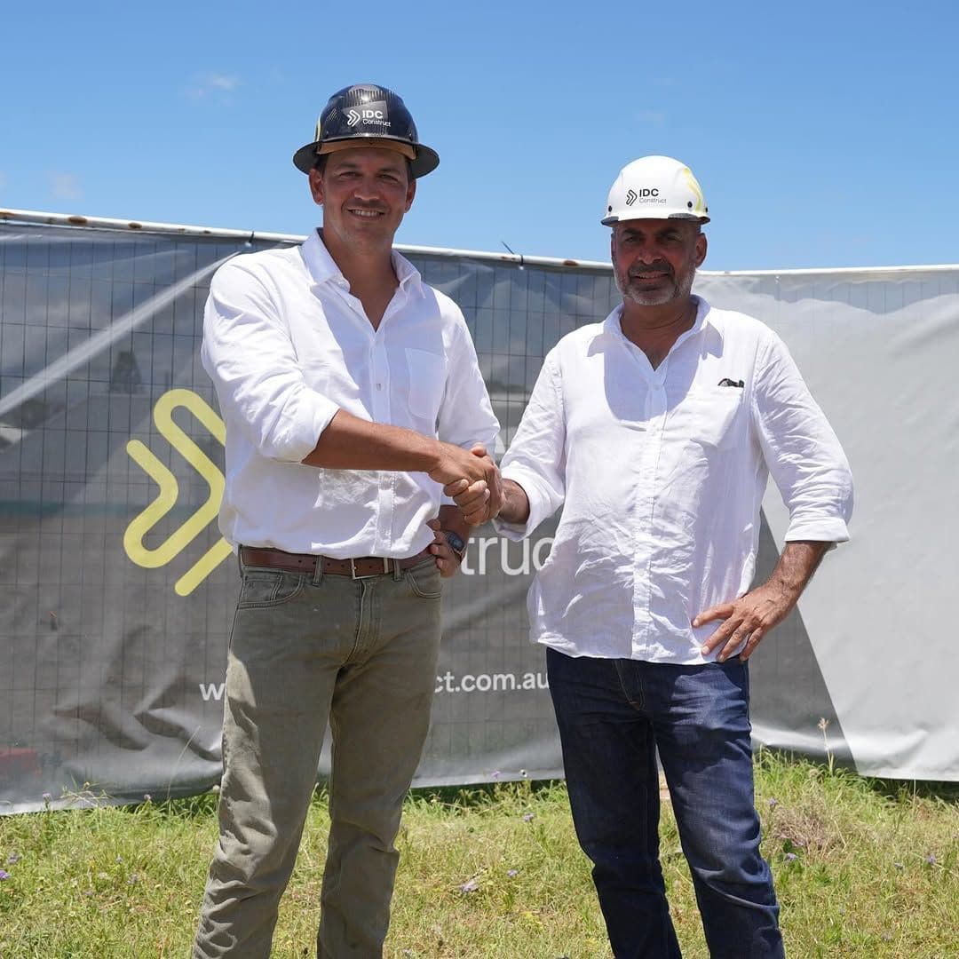 Two men in white shirts and IDC Construct hard hats shaking hands in front of the site hoarding at the start of construction